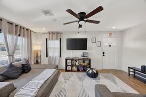 Una sala de estar moderna con un sofá, un ventilador de techo, un televisor de pantalla plana, una estantería y una puerta señalizada con 'EXIT'.