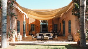 A covered outdoor seating area with a sofa and chairs, palm trees, and a building with arched windows. - Villa-Riad with Private Pool in Marrakech, Akhdar 1 (Marrakech)