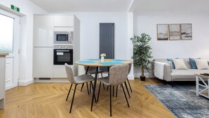 A modern living room with a sofa, a dining table with chairs, a potted plant, and a kitchen area with appliances. - Entire Home Next to Trafford Centre (Stretford)