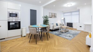A modern kitchen with built-in appliances, a dining area with a round table and chairs, a sofa, and a large window with blinds. - Entire Home Next to Trafford Centre (Stretford)