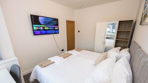 A neatly made bed with white linens, a television mounted on the wall displaying a home screen, a wardrobe with a mirror, and a small desk with a chair. - Entire Home Next to Trafford Centre (Stretford)