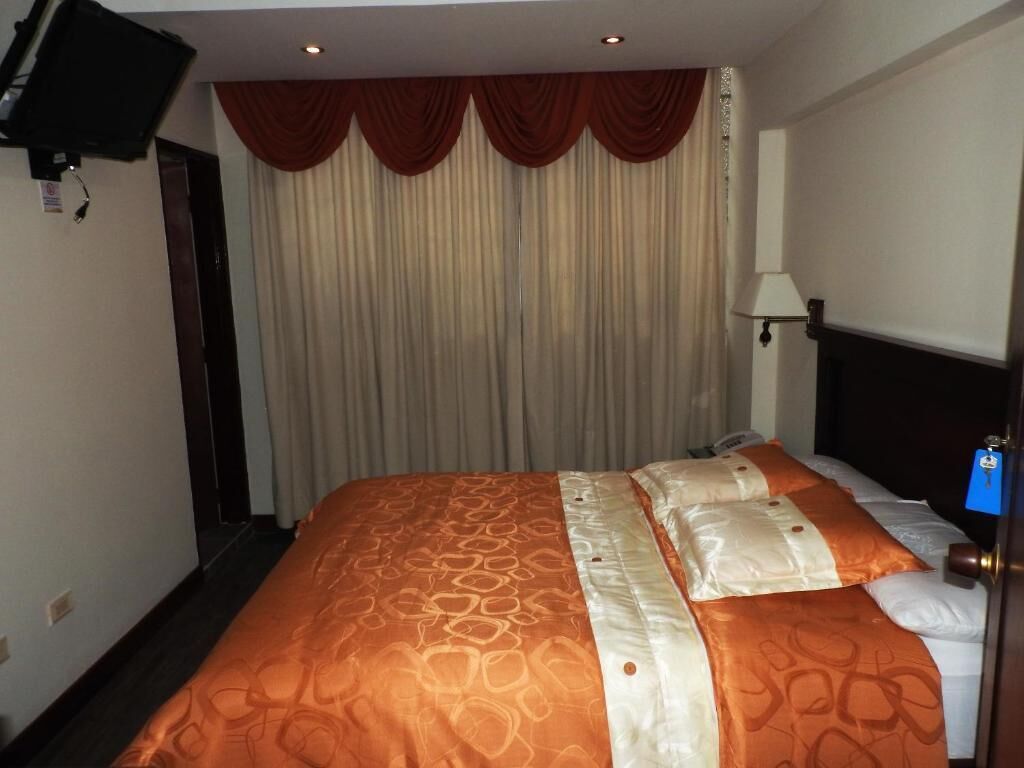 Una habitación de hotel con una cama, cortinas, un televisor montado en la pared y una lámpara.