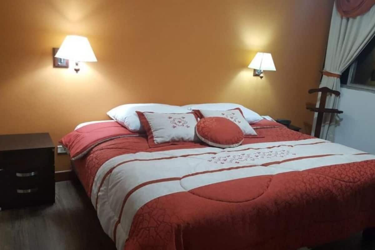 A bed with a red and white bedspread, two wall-mounted lamps, a nightstand, and a window with curtains.