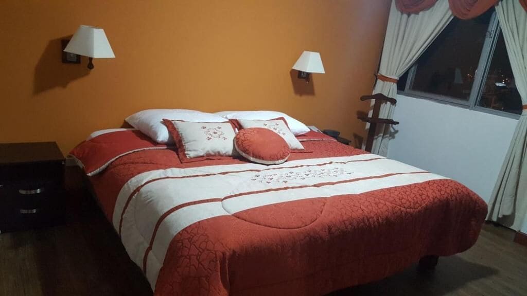 Un lit recouvert d’une couvre-lit rouge et blanche, deux lampes fixées au mur et une fenêtre avec des rideaux.