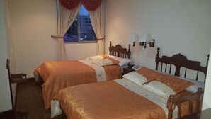A hotel room with two beds, a nightstand, a lamp, and a window with curtains. - Hotel Angasmayo (Ipiales)