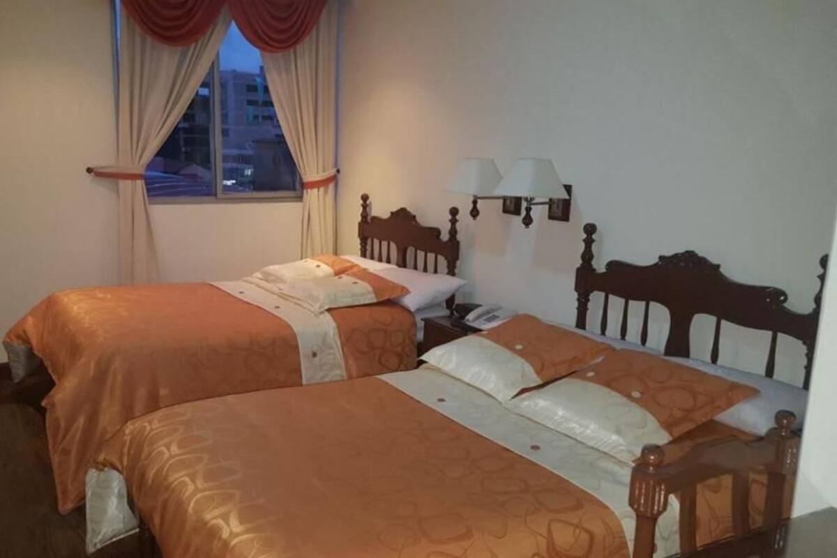 A hotel room with two beds, a nightstand, a lamp, and a window with curtains.