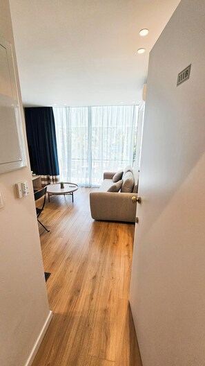 A modern living room with a sofa, a small round table, and a chair. There is a large window with sheer curtains, and a door with a number plate. - The Colors of Larnaca (Larnaca)