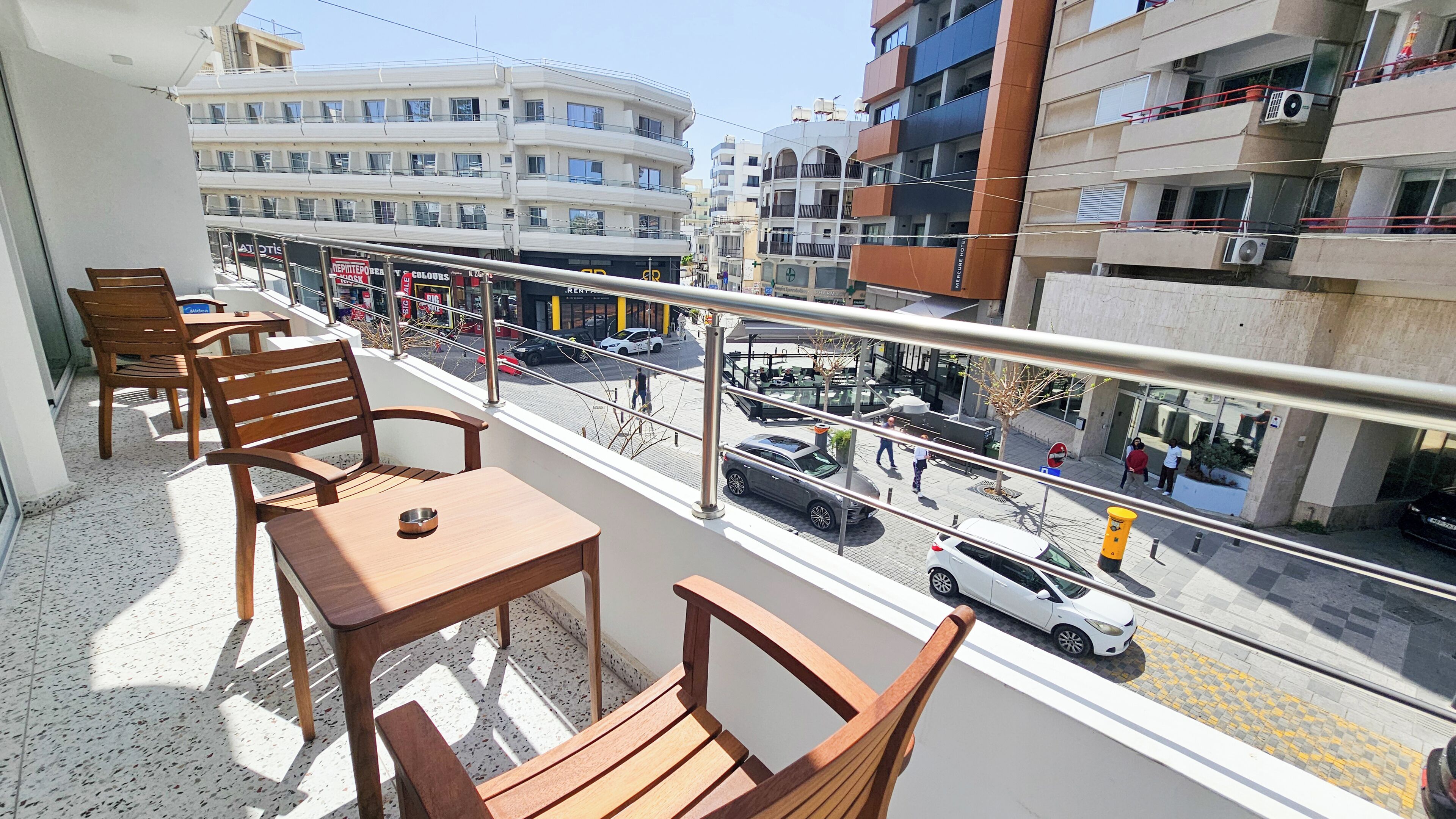 Un balcon avec des chaises et une table en bois, donnant sur une rue animée où circulent des voitures et des piétons.