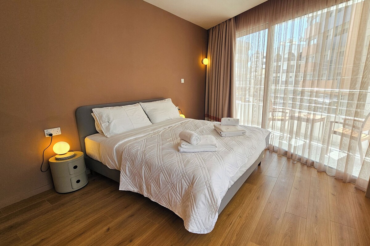 A hotel room with a bed, a bedside lamp, a nightstand, and a window with curtains.