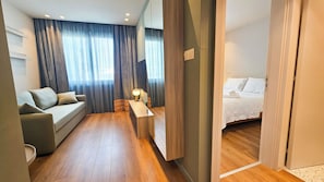 A modern hotel room with a sofa, a desk with a lamp, a bed, and a view through a window with curtains. - The Colors of Larnaca (Larnaca)