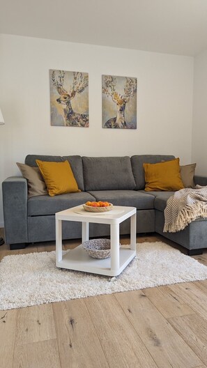 Un salon avec un canapé gris, deux coussins de couleur moutarde, une table basse blanche avec un bol, et deux tableaux représentant des cerfs accrochés au mur.