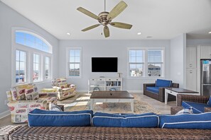 A spacious living room with a ceiling fan, large windows, a flat-screen TV, and a dining area with wicker furniture. - The Caddyshack at Carolina Beach (Carolina Beach)