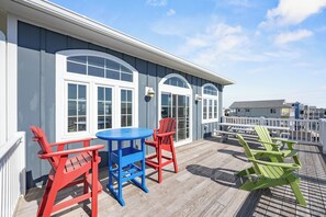 Une terrasse avec une table bleue et des chaises Adirondack rouges et vertes, une balustrade blanche et un bâtiment doté de grandes fenêtres.