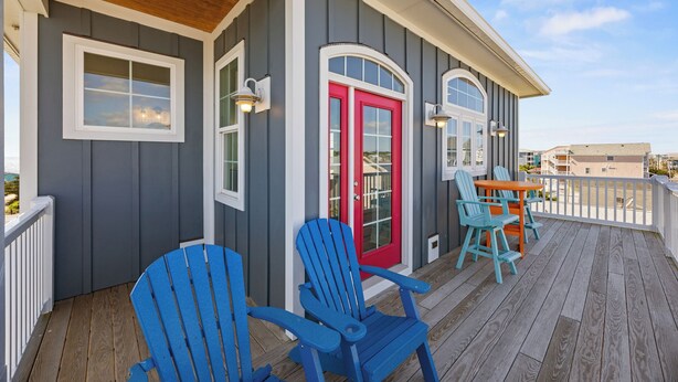 Eine überdachte Veranda mit blauen Adirondack-Stühlen, einer roten Tür und einem weißen Geländer.