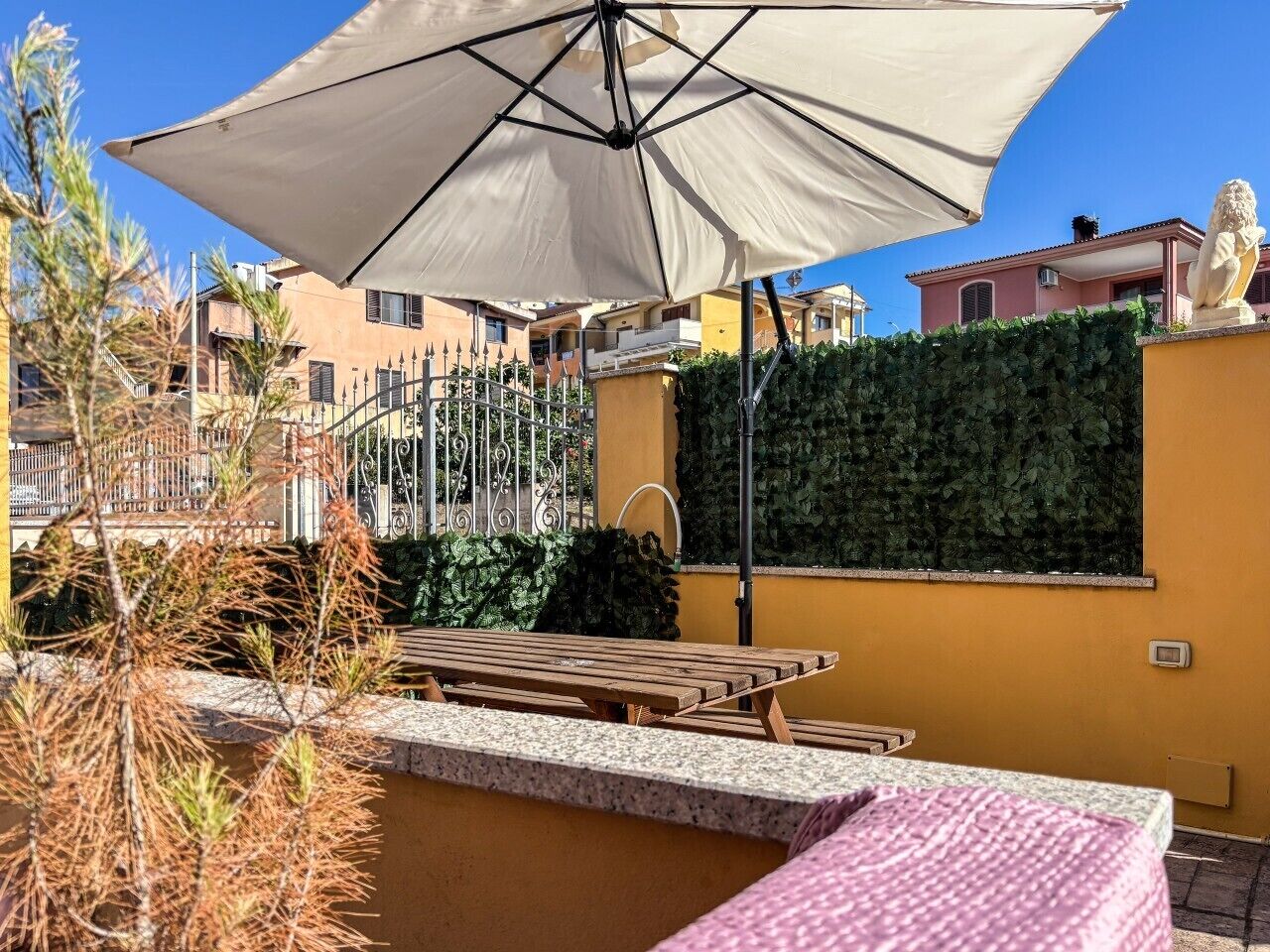 Un patio con tavolo e ombrellone, un siepe e un edificio sullo sfondo.