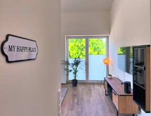Ein modernes Interieur mit einem Schild mit der Aufschrift 'MY HAPPY PLACE', einem Holztisch, einem Fernseher und einem großen Fenster mit Blick auf Grünflächen.