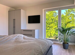 Ein Schlafzimmer mit Bett, Kleiderschrank, Flachbildfernseher und einem großen Fenster mit Blick auf Bäume.