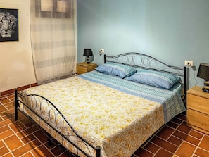 Un dormitorio con estructura metálica para la cama, colcha estampada con motivos florales, dos almohadas, una mesita de noche con lámpara y un cuadro colgado en la pared.