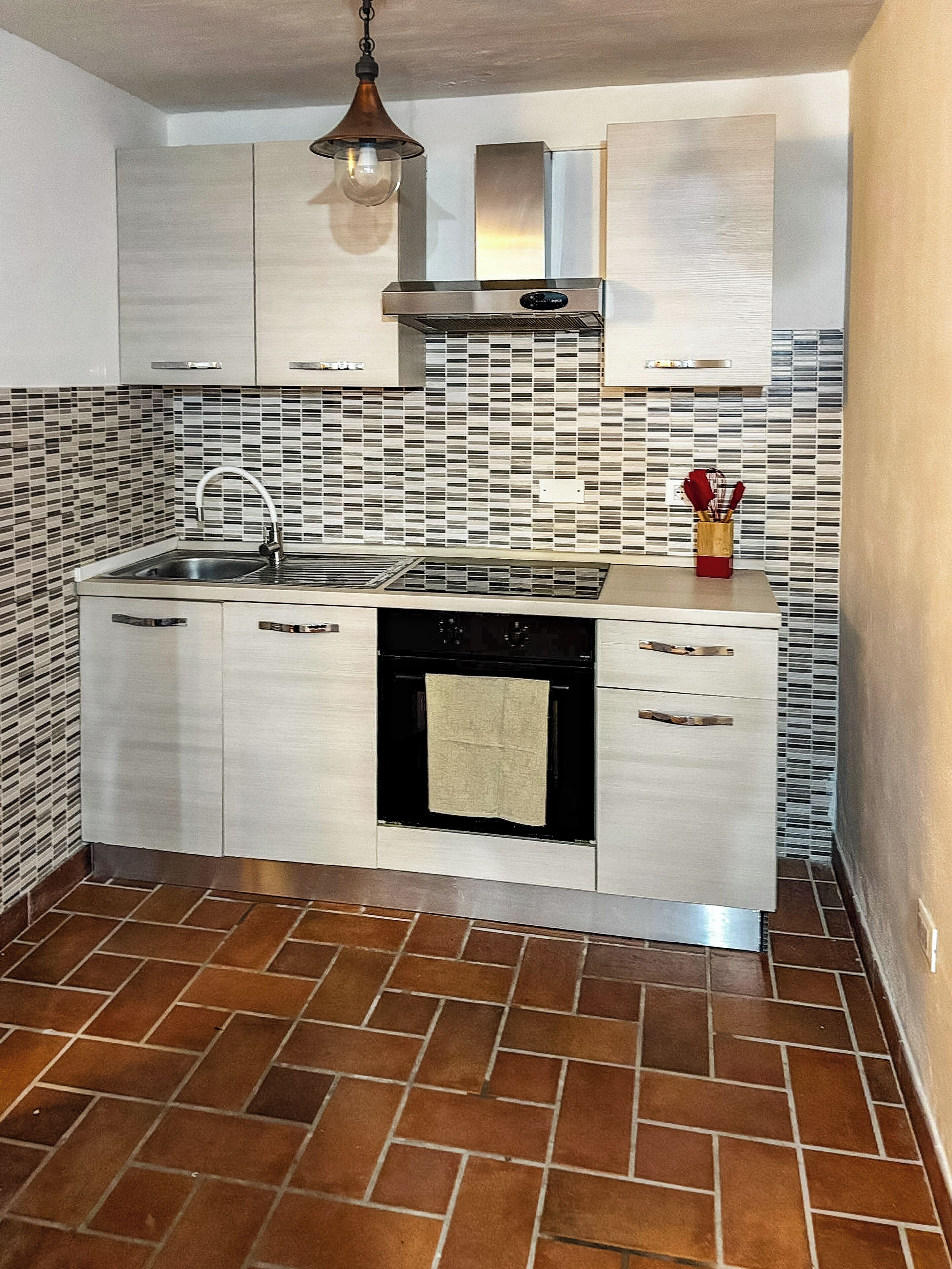 A kitchen with a tiled backsplash, a stainless steel range hood, a built-in oven, and a hanging light fixture.