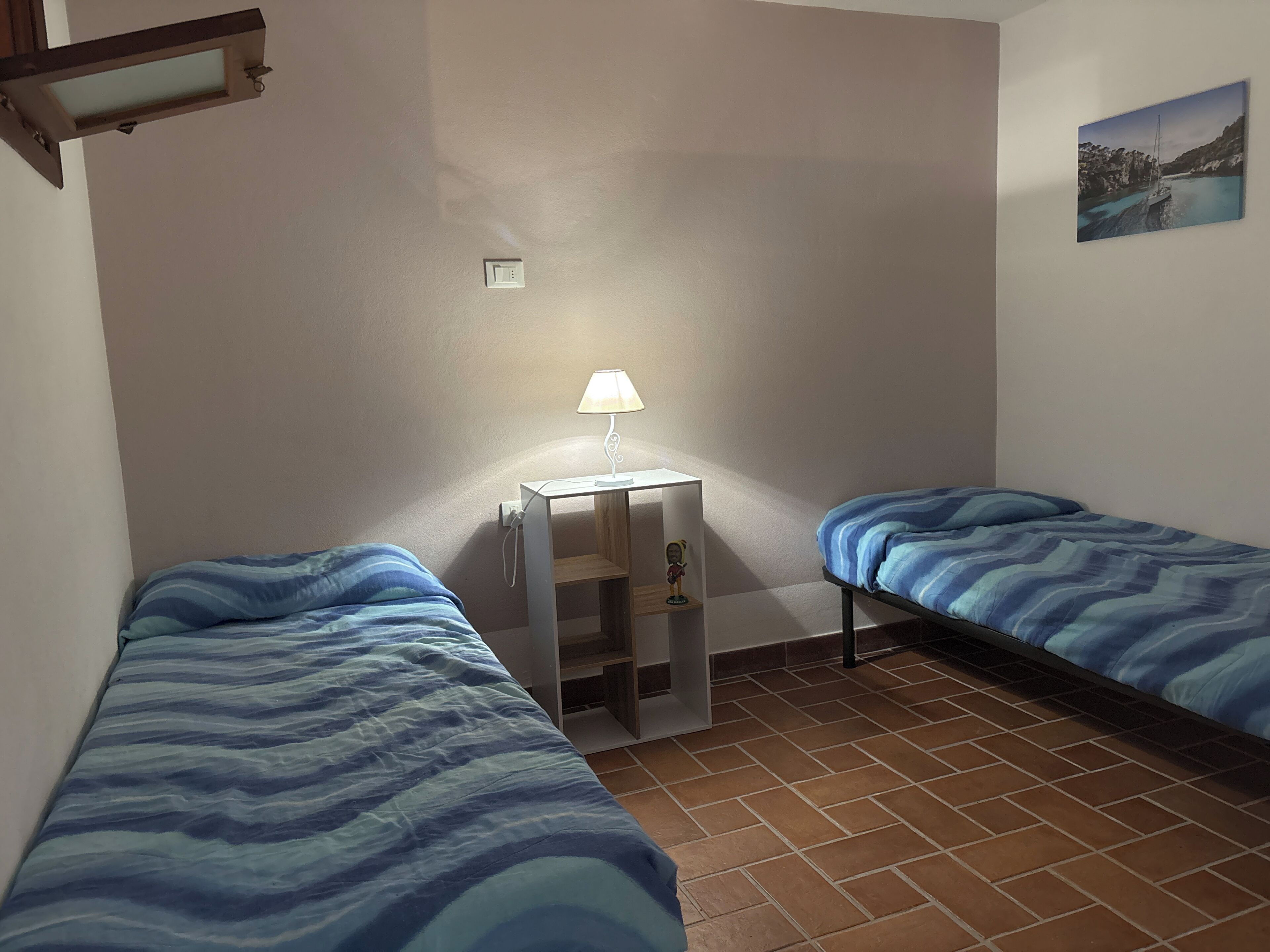Ein Zimmer mit zwei Betten, einem Nachttisch mit Lampe, einem kleinen Regal mit einer Figur und einem Bild an der Wand.