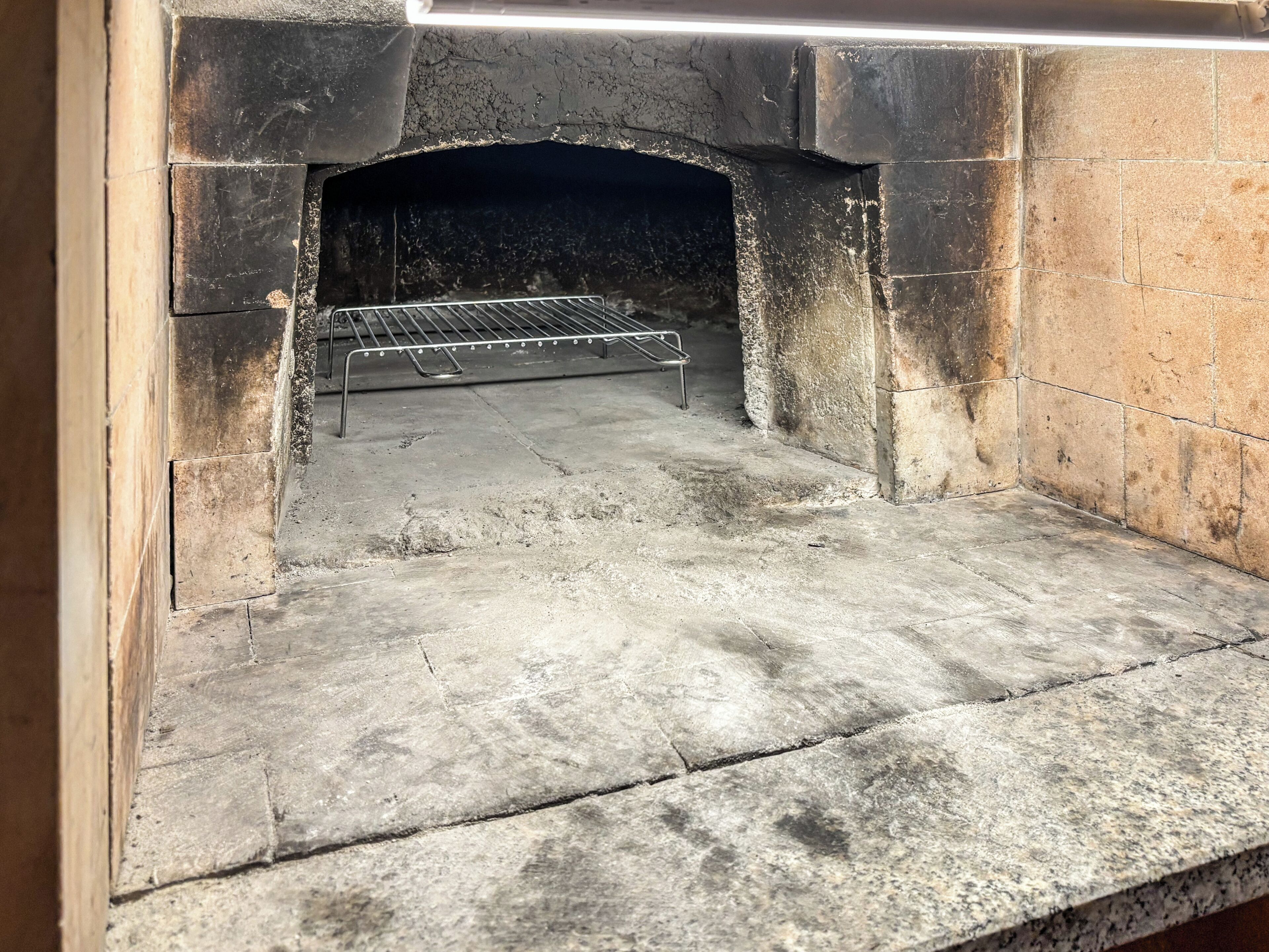 Un horno de piedra con una rejilla metálica en su interior, suelo de concreto y pared de ladrillo.