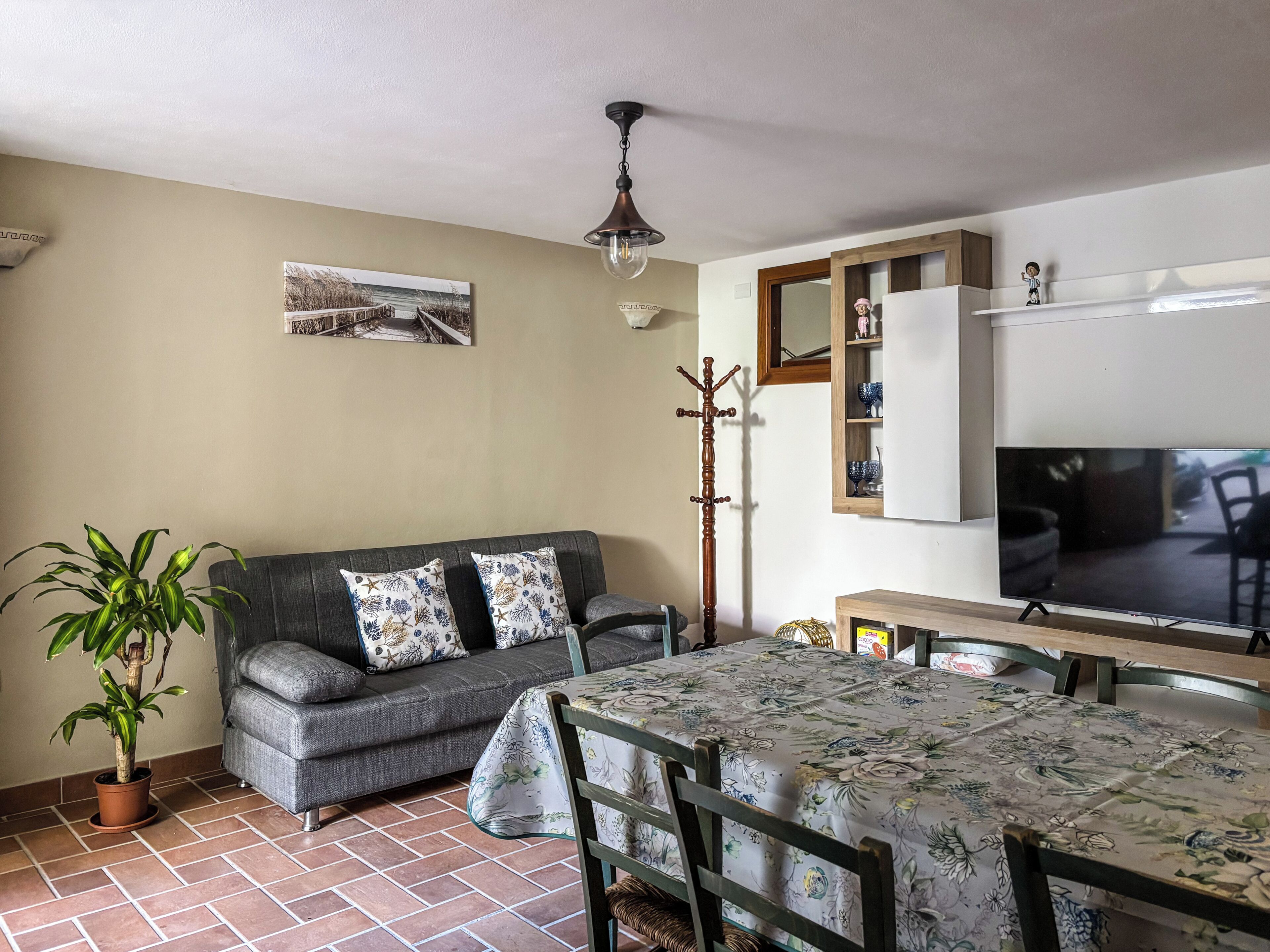Uma sala de estar com sofá, mesa de jantar, televisão, cabideiro e planta em vaso.