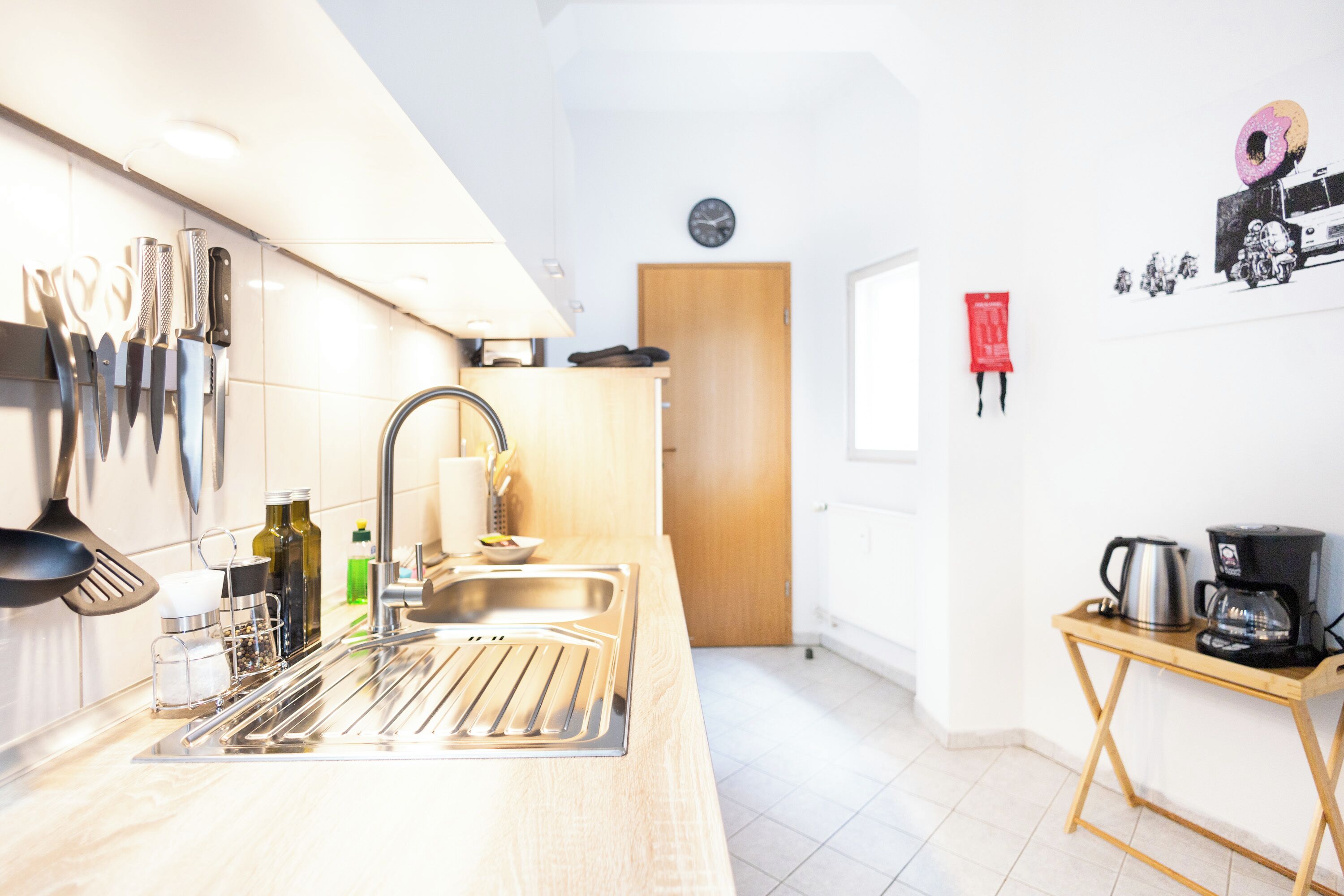 A modern kitchen with a stainless steel sink, hanging knives, a wooden door, and a small table with a coffee maker.