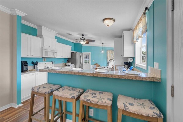 A kitchen with white cabinets, a central island with stools, a ceiling fan, and a window with curtains. - Fish, Boat & Relax-Direct Access Canal Home Near Top Fishing Spots (Punta Gorda)