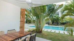 Un patio con tavolo e sedie in legno, una ventola a soffitto e vista su una piscina circondata da piante tropicali.