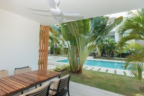 A patio with a wooden table and chairs, a ceiling fan, and a view of a swimming pool surrounded by tropical plants. - Pool View and 2 min from the beach: Condo in El Cortecito (Punta Cana)