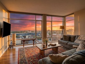 A spacious living room with a large window, a flat-screen TV, a sofa, a coffee table, and a lamp. - Lakeview Condo + Balcony + Parking + Gym (Cleveland)