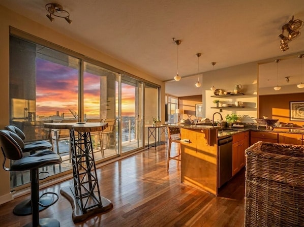 A modern kitchen with a large island, bar stools, and a view of a sunset through floor-to-ceiling windows. - Lakeview Condo + Balcony + Parking + Gym (Cleveland)