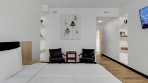Une chambre d’hôtel moderne avec un grand lit, deux fauteuils, une petite table, un tableau au mur et un téléviseur sur le côté droit.