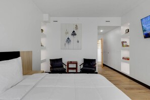 Ein modernes Hotelzimmer mit einem großen Bett, zwei Sesseln, einem kleinen Tisch, einem Bild an der Wand und einem Fernseher auf der rechten Seite.