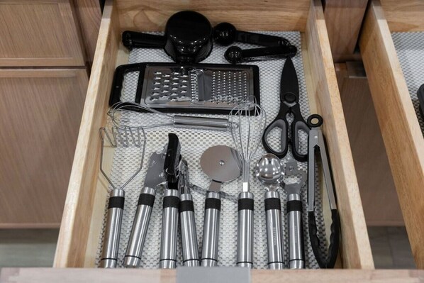 Un cajón con utensilios de cocina, incluyendo un rallador, batidor, tijeras y varias herramientas metálicas.