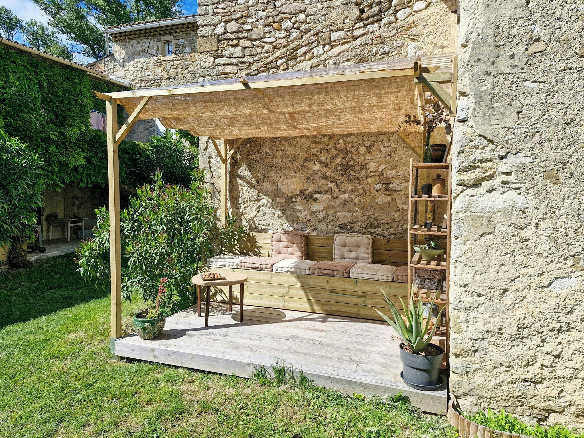 A stone building with a wooden porch, a seating area with cushions, and a small table with a potted plant.