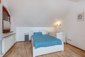Une chambre avec un lit blanc, un radiateur, une fenêtre avec des stores, une lampe fixée au mur et une petite armoire blanche.