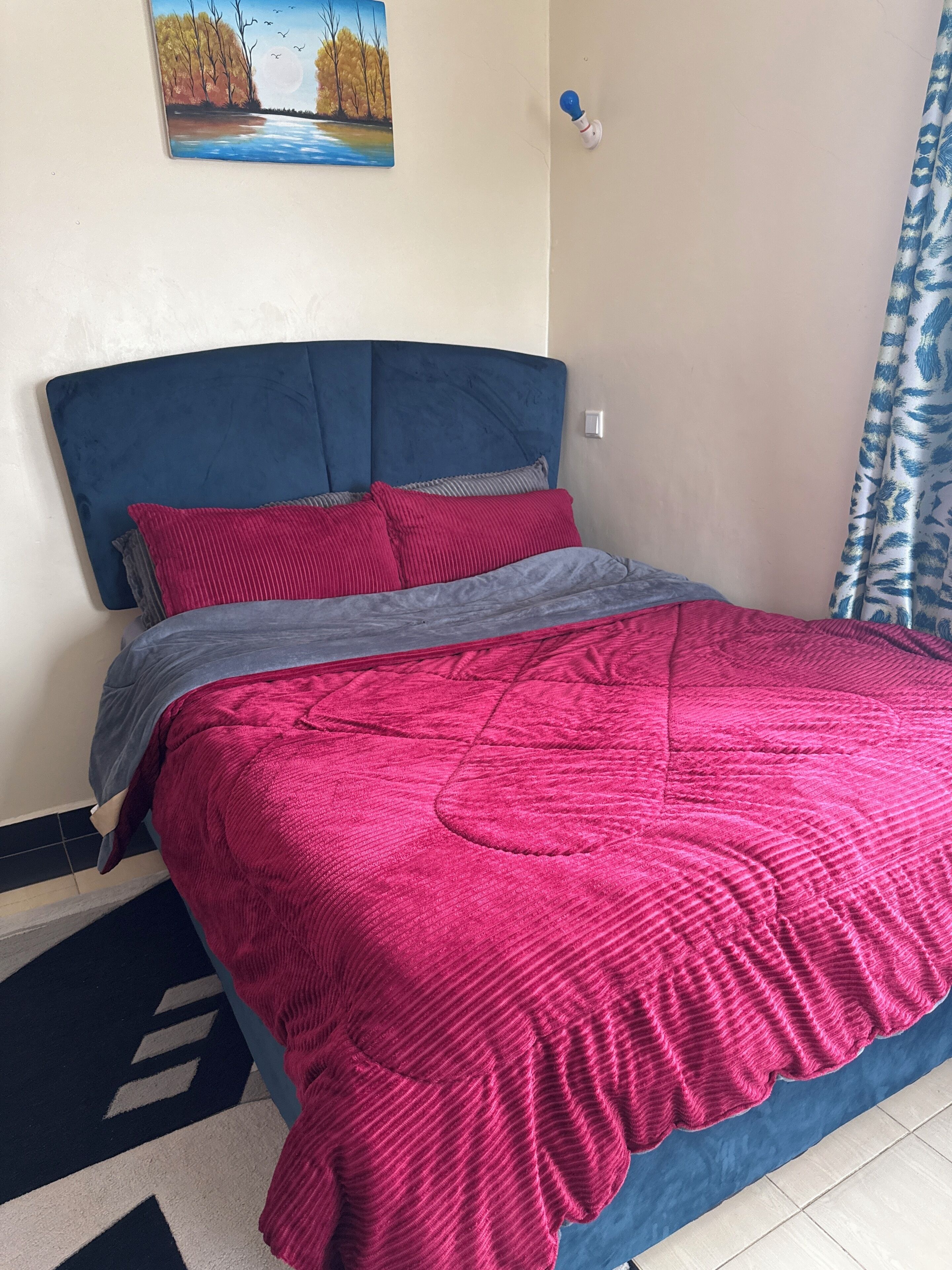 Een bed met een blauw hoofdeinde, rode en grijze beddengoed, en een schilderij van een landschap aan de muur.