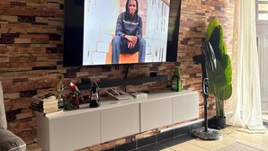 A living room with a television on a brick-patterned wall, a white entertainment unit, a large green plant, and a grey sofa. - Bright 2-bedroom apartment with AC in serene Nairobi (Nairobi)