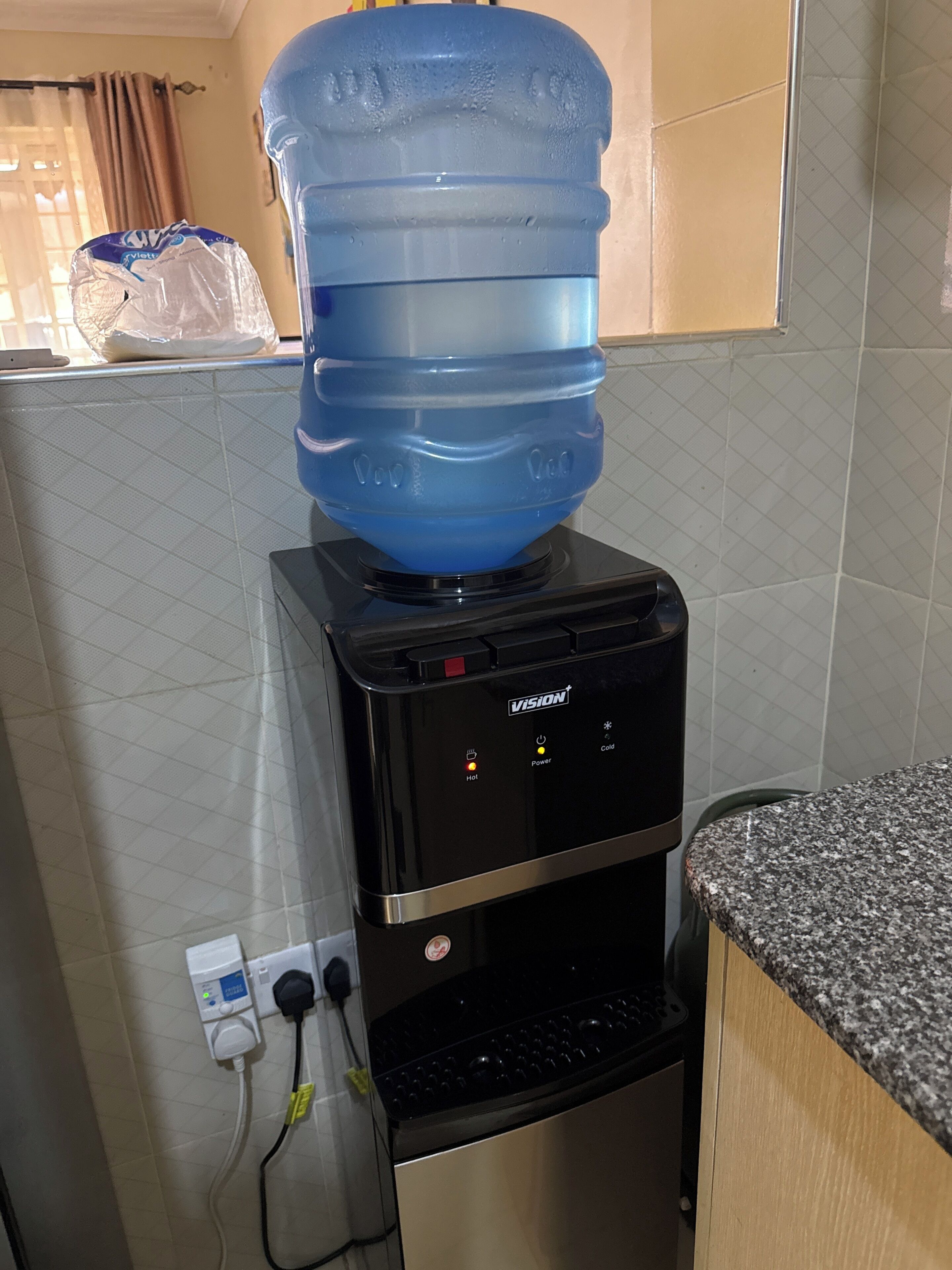 Een waterdispenser met een grote blauwe waterkan op de bovenkant, een bedieningspaneel met warm, power en koud instellingen, en een granieten aanrecht.