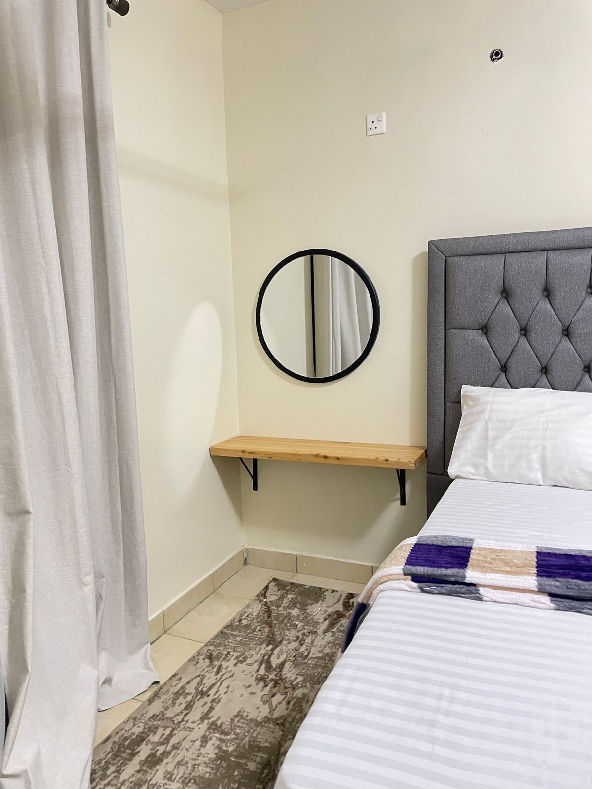 A bedroom with a bed, a round mirror, a wooden shelf, and a patterned rug.