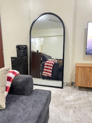 A living room with a grey sofa, a coffee machine, a wooden cabinet, and a large mirror reflecting the room. - JB VACATION HOME (Mombasa)