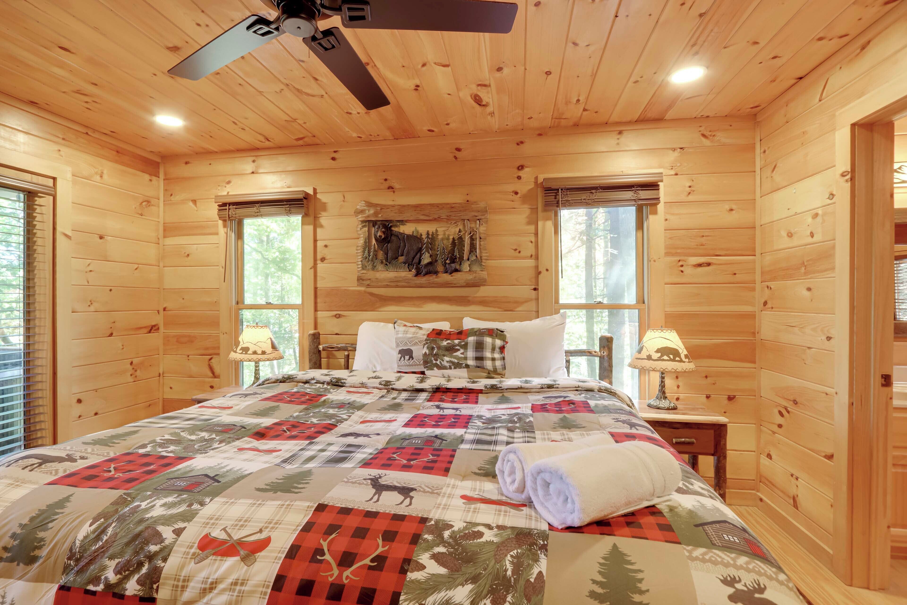 A wooden cabin bedroom with a bed, two nightstands, a ceiling fan, and a window with a view of trees.