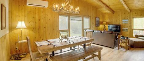 A wooden interior with a dining table, benches, a chandelier, a television, and a kitchen area.