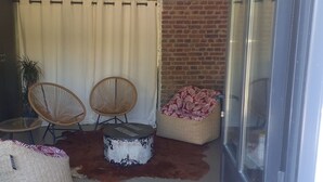 Una habitación con puerta de vidrio, dos sillas de mimbre, un otoman redondo y una mesita, además de un otoman con estampado.