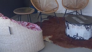 Una habitación con un sofá, dos sillones de mimbre, un otoman redondo, un armario oscuro y una cortina.