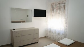 Un dormitorio con una cama, un aparador, un televisor y una ventana con cortinas translúcidas.