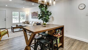 Uma área de refeições com mesa de madeira, banquetas de bar, luminária pendente, planta em vaso e relógio na parede.