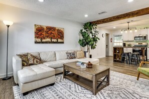 A living room with a sectional sofa, a coffee table, a painting of trees, a large plant, and a kitchen in the background. - < 2 Mi to Dallas Stadium! Work & Play Base w/ Yard (Arlington)