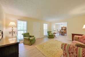 A living room with a sofa, chairs, a rug, a lamp, and a dining area in the background.